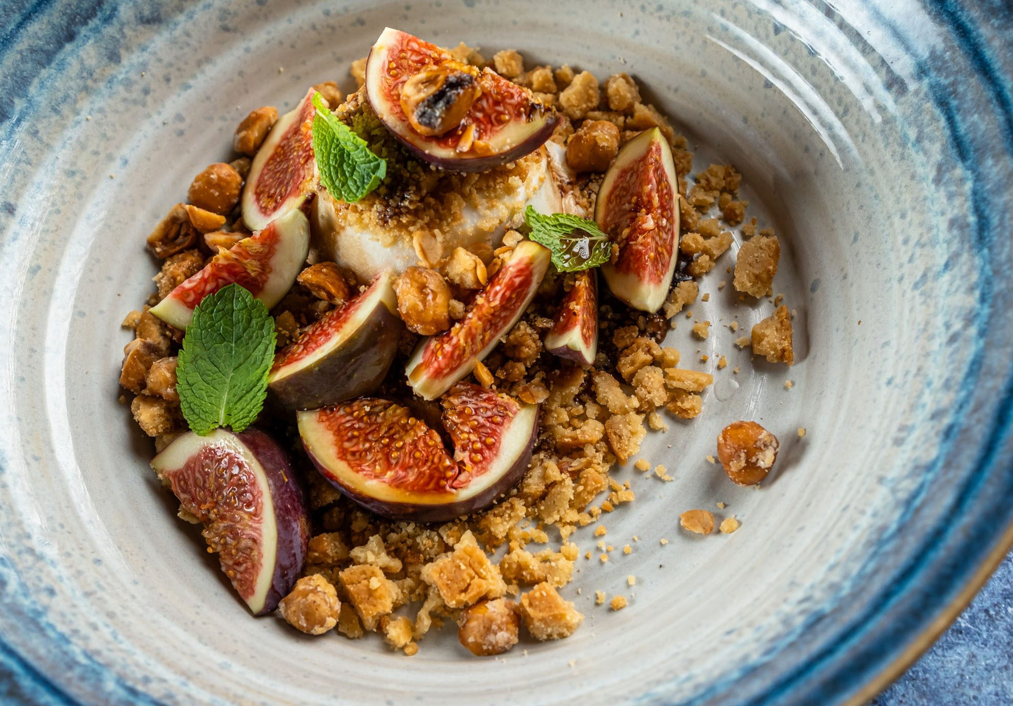 Gourmet étel, amely krémes panna cottát tartalmaz, szeletelt fügével és mentalevelekkel tálalva, bézs színű tányéron, morzsolt dióval és keksszel körítve.